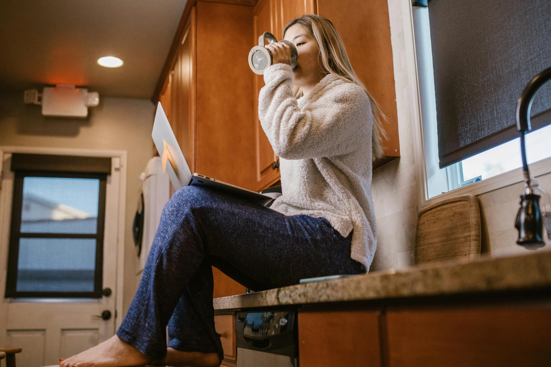 Femme travaillant depuis sa cuisine avec un ordinateur et une tasse en main.